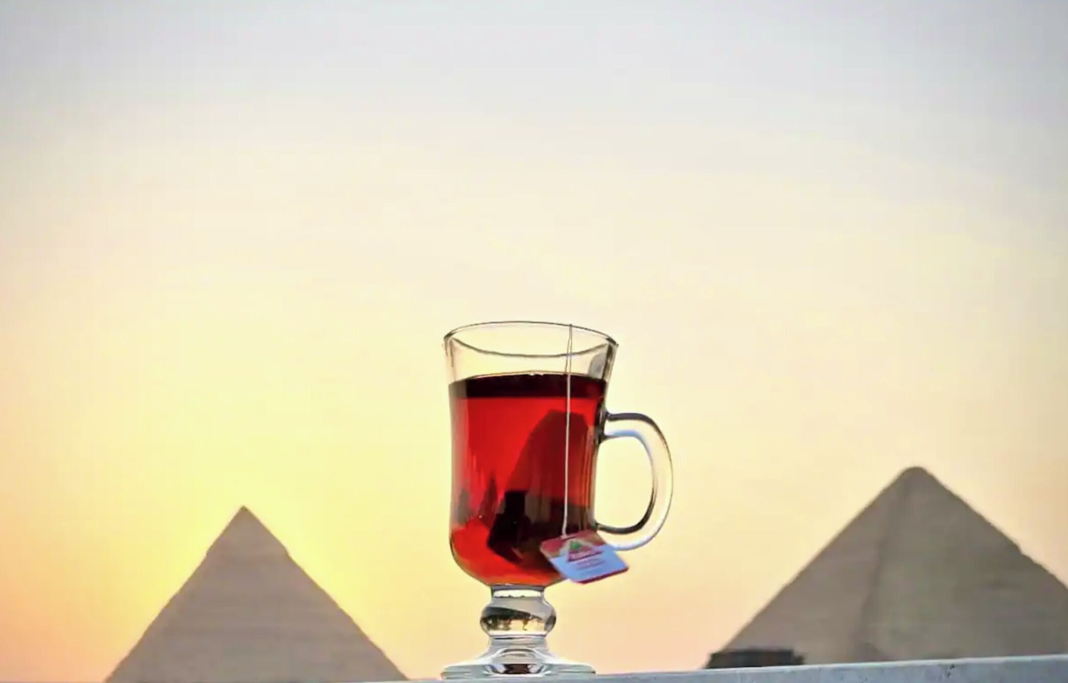 A clear glass mug with a tea bag, placed on a surface with the pyramids in the background.