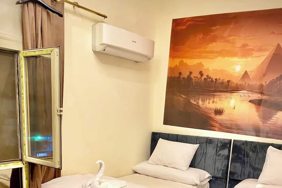 A hotel room with two beds, a large window, an air conditioning unit, and a sunset painting on the wall.