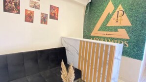 Uma área de recepção com um sofá preto, uma mesa com tampo de vidro e um vaso, e uma parede verde com um logotipo.