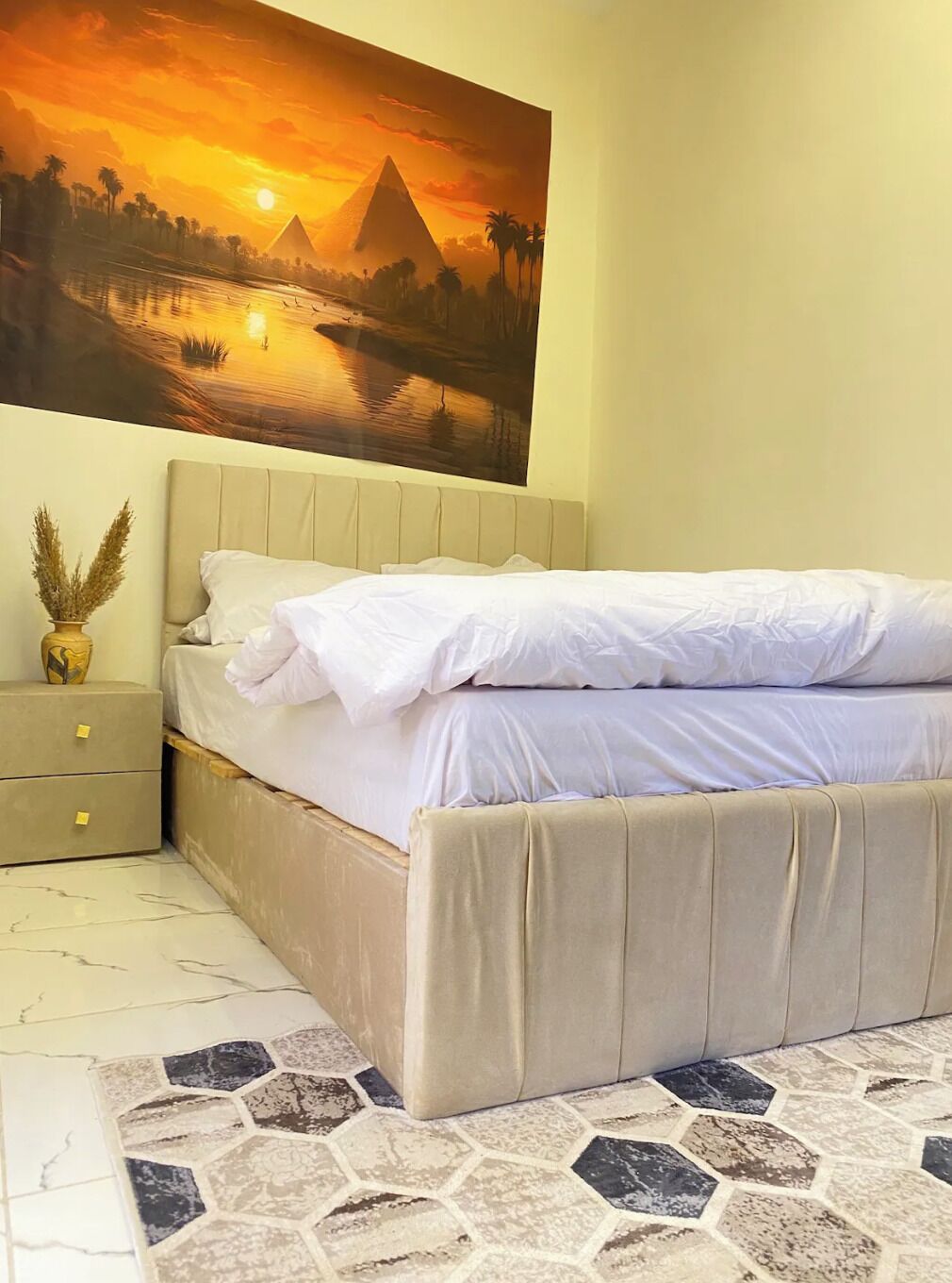 Ein Schlafzimmer mit einem Bett, einem Nachttisch, einer Vase mit Trockenpflanzen und einem Gemälde, das einen Sonnenuntergang über einer Pyramide zeigt.