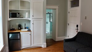 Une cuisine compacte avec des meubles blancs, un four à micro-ondes, une machine à café et un réfrigérateur. Le salon est équipé d’un canapé de couleur foncée.