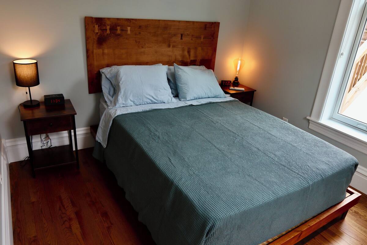 A bedroom with a wooden headboard, a bed with a green bedspread, two bedside lamps, and a nightstand with a clock.