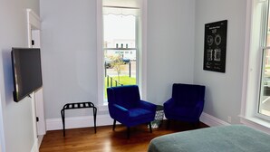 Une pièce avec deux fauteuils bleus, un lit, une télévision et une fenêtre donnant sur un immeuble et un arbre.