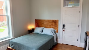 Une chambre à coucher avec une tête de lit en bois, un lit recouvert d’une couverture verte, une table de chevet avec une lampe et une porte blanche portant un numéro au-dessus.