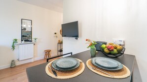 A modern dining area with a table set for two, a fruit bowl, a vase with flowers, and a mounted TV.