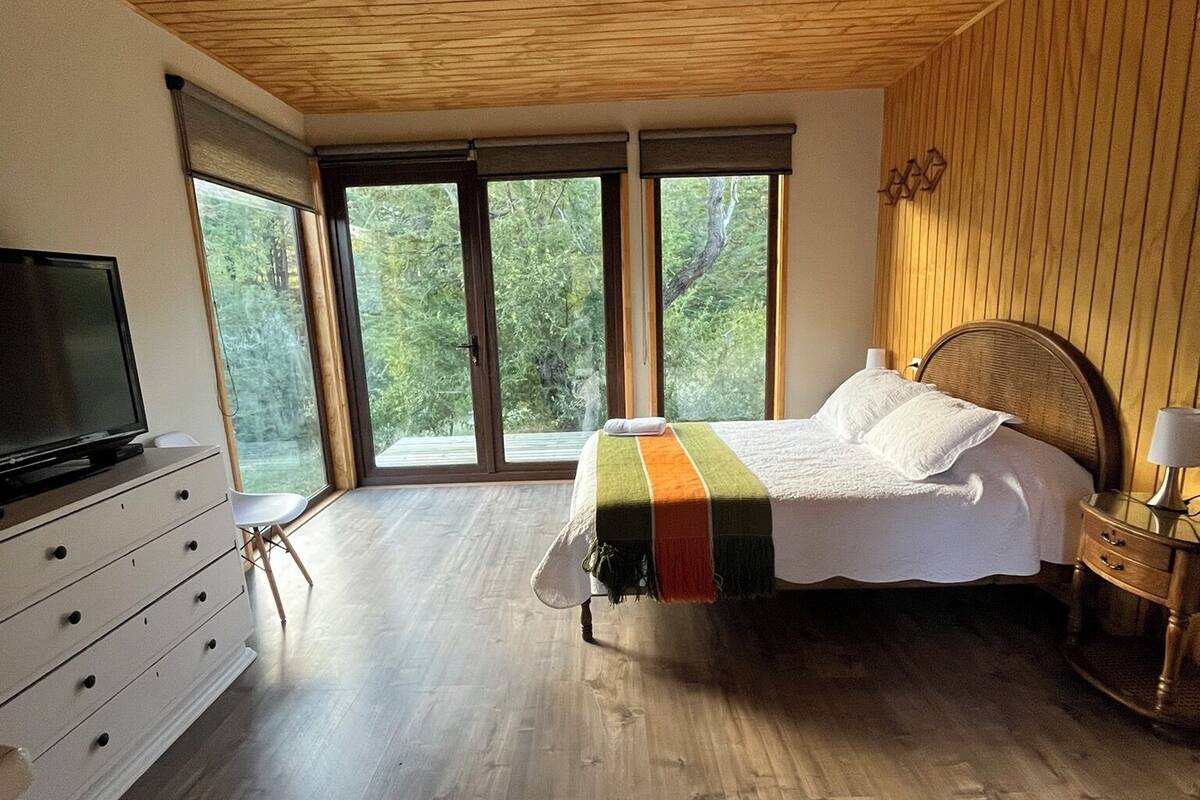Een slaapkamer met een houten plafond, een bed met een kleurrijke deken, een televisie op een dressoir en grote ramen met uitzicht op bomen.