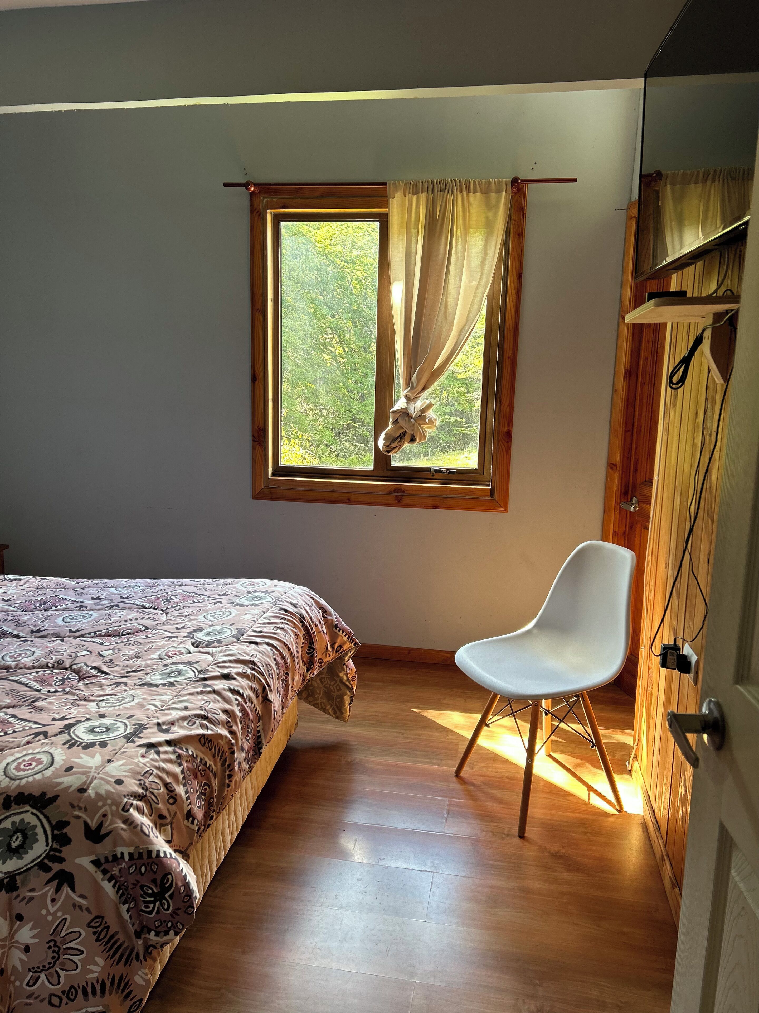 Un dormitorio con cama, una silla, un ventanal con cortinas y paredes de madera.