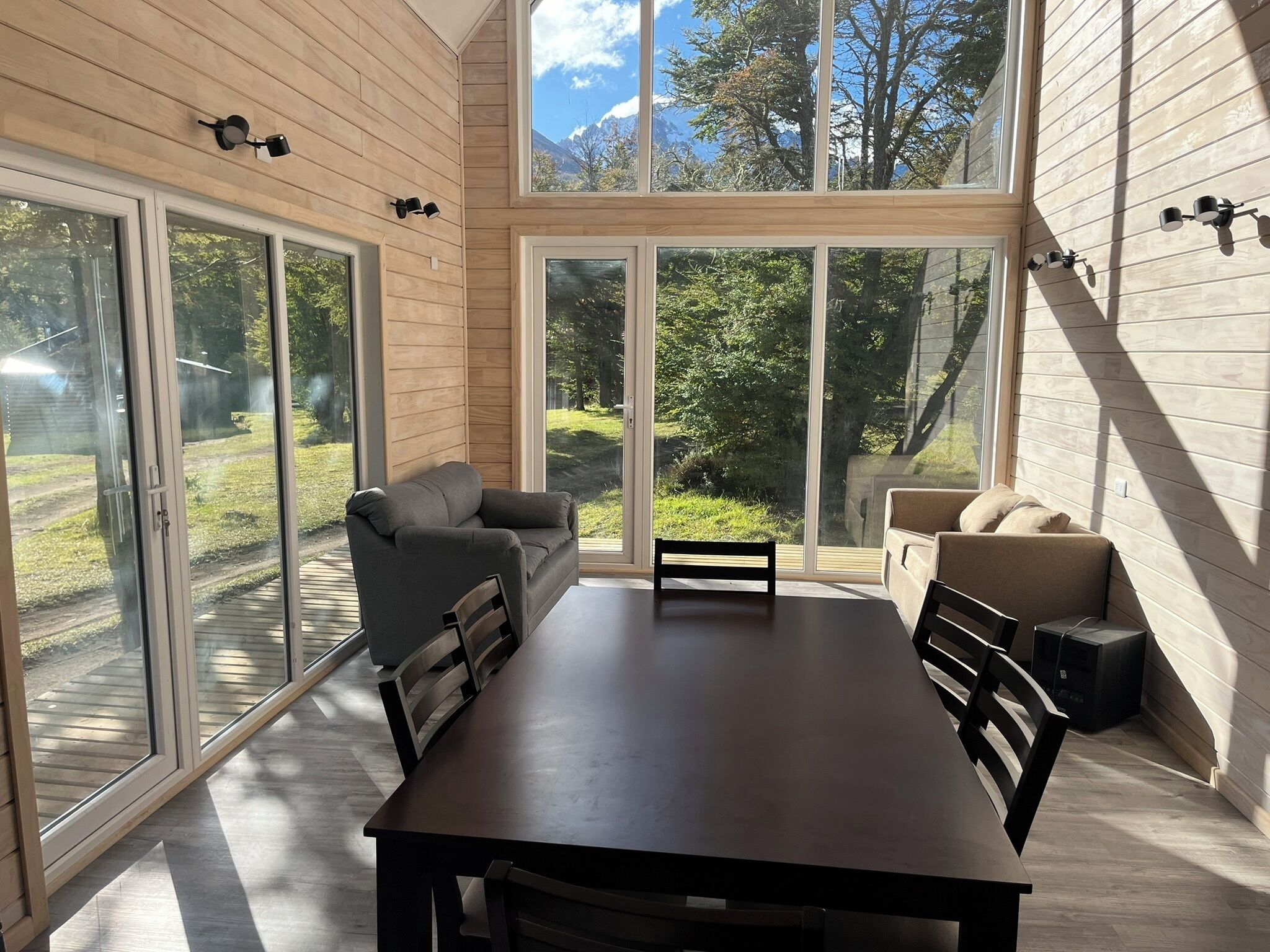 Un interior moderno con una mesa de comedor, dos sofás y amplias ventanas con vistas a un jardín y montañas.