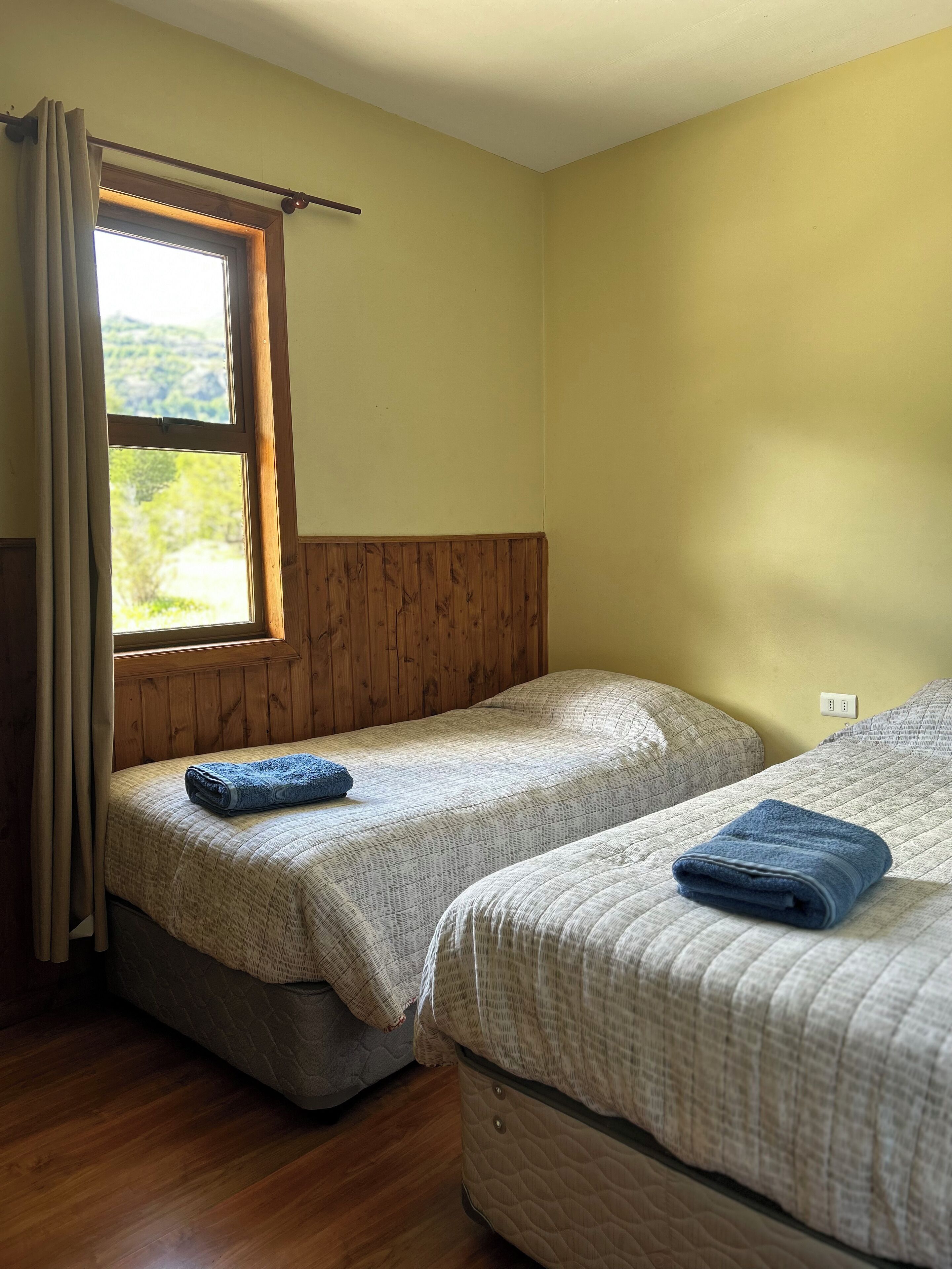 Een kamer met twee bedden, houten hoofdeinden, een raam met gordijnen en uitzicht op groen.