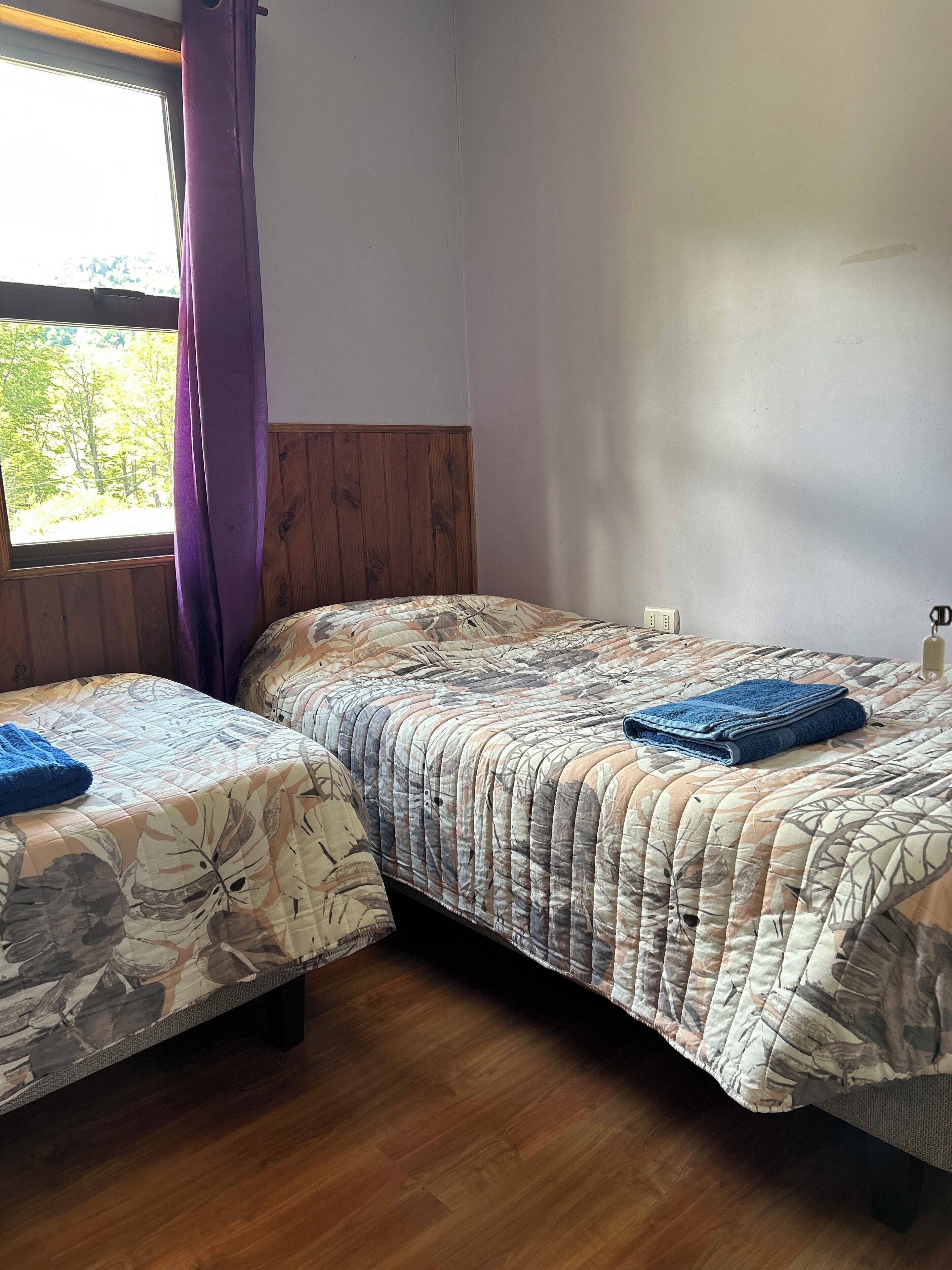 A room with two beds, wooden headboard, a window with curtains, and a view of trees outside.