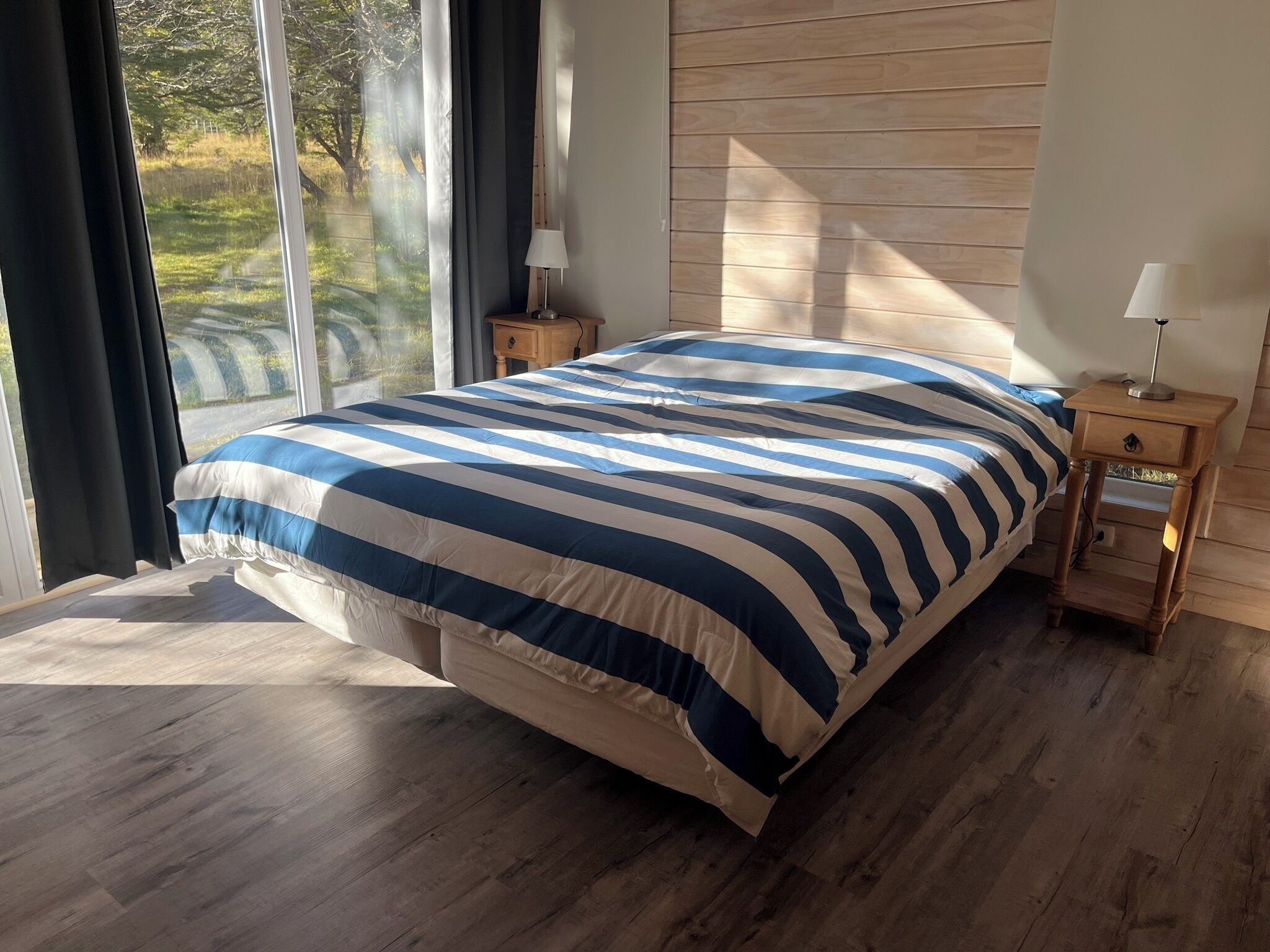 Quarto com uma cama grande, piso de madeira, mesa de cabeceira com abajur e vista para o verde pela janela.