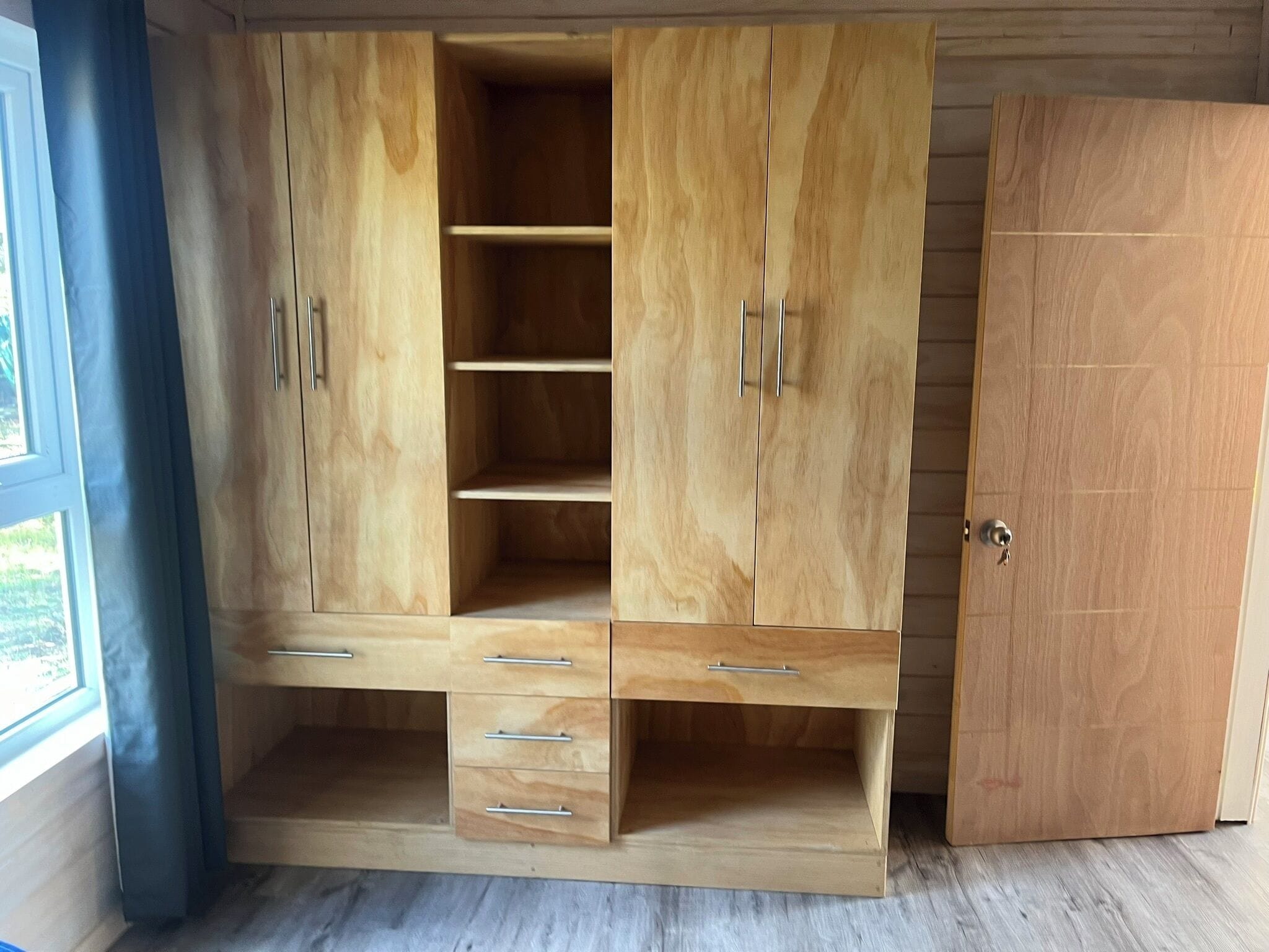 A wooden wardrobe with multiple compartments and drawers, a door, and a window with curtains.