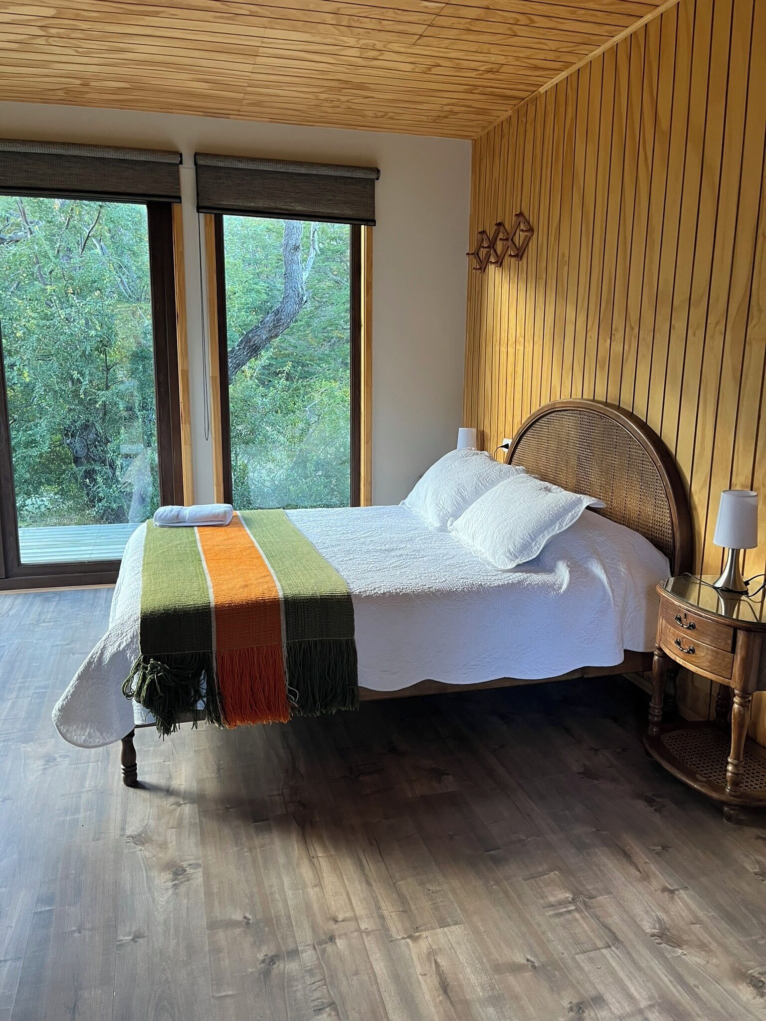 A bedroom with a large window, a bed with a colorful blanket, a wooden headboard, and a bedside table with a lamp.