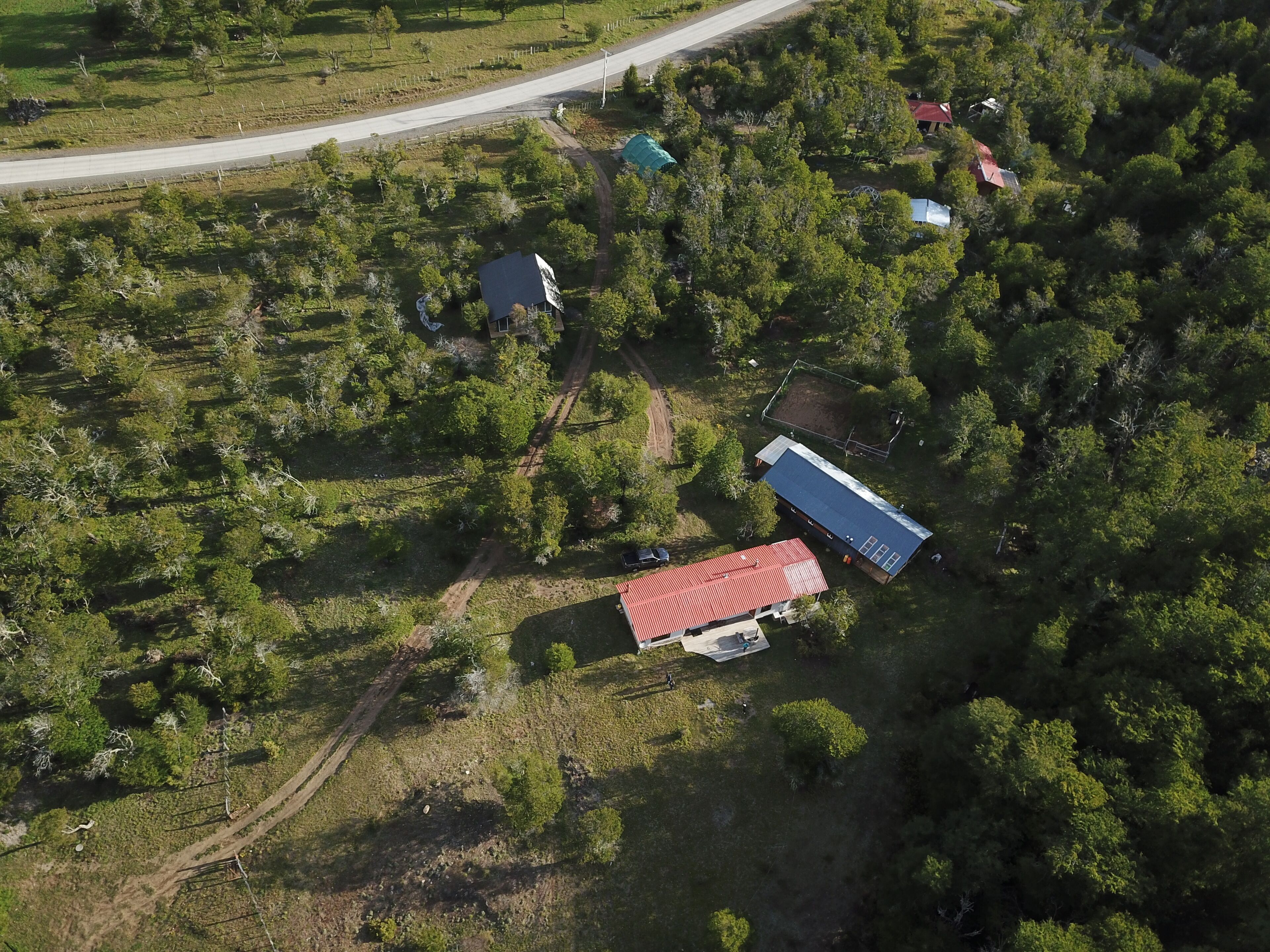 An aerial view of a rural area with a main building featuring a red roof, surrounded by dense greenery and a few smaller structures.