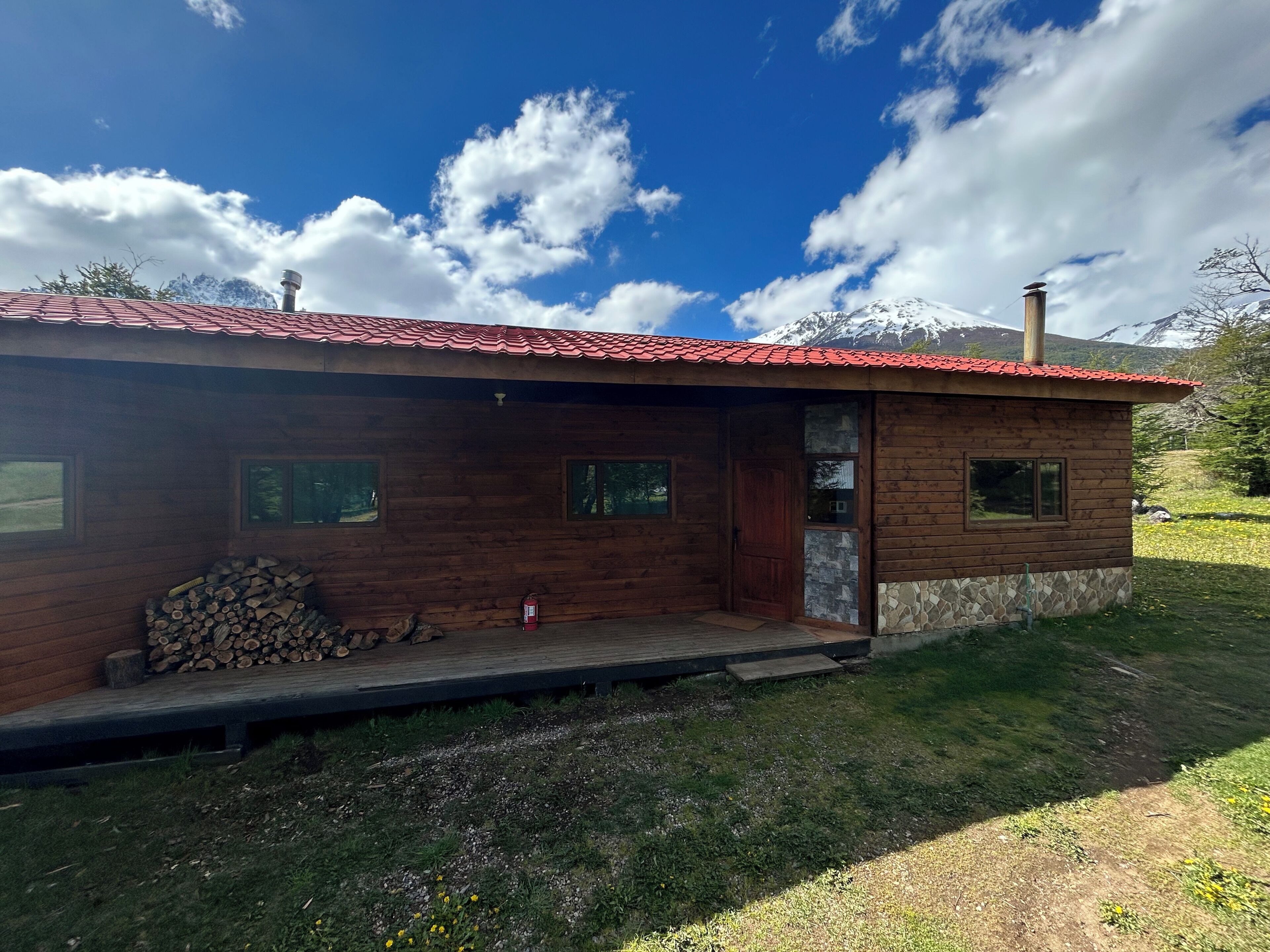 Une cabane en bois avec un toit recouvert de tuiles rouges, une véranda en bois et une vue sur des montagnes enneigées en arrière-plan.