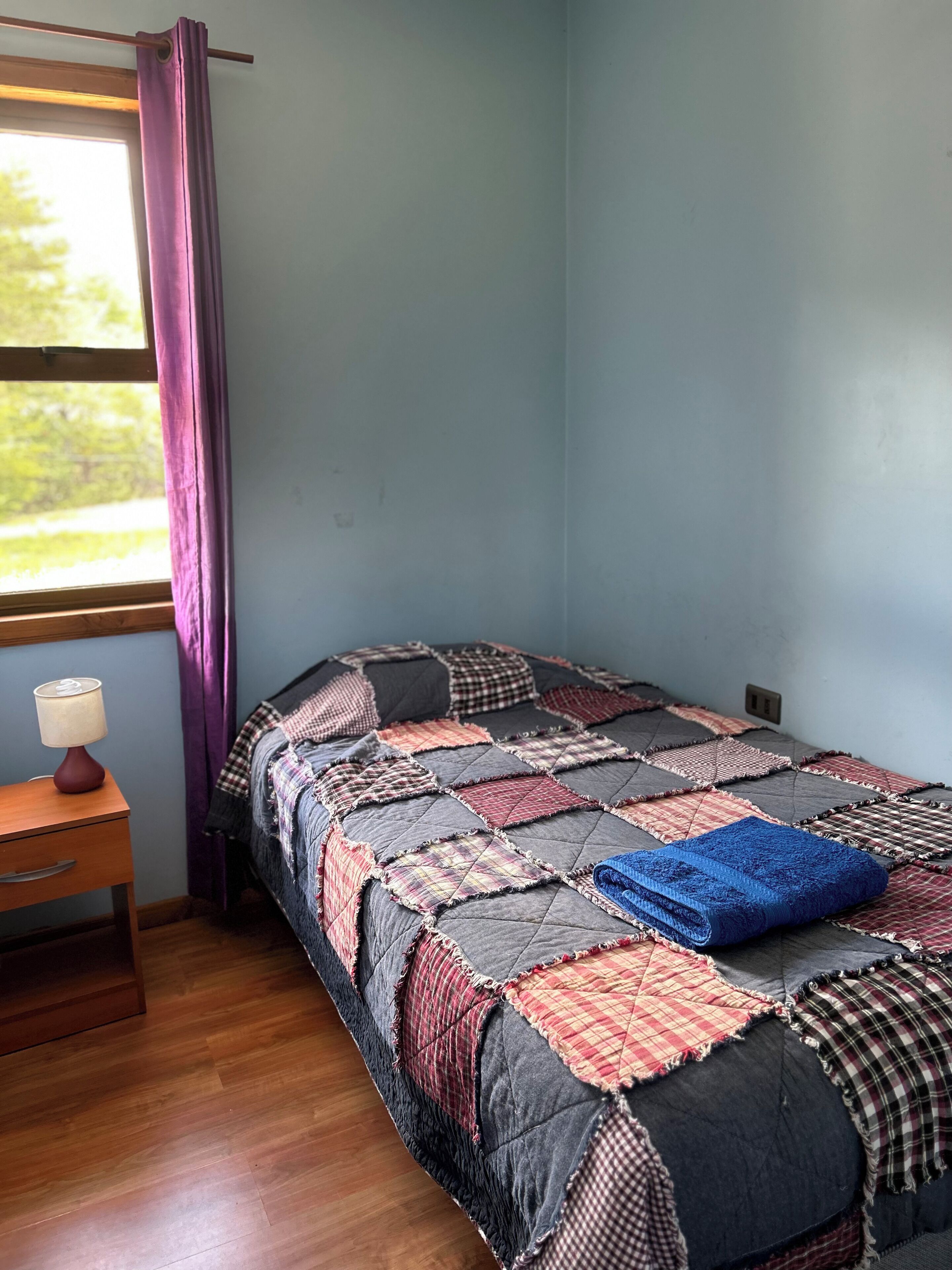 A single bed with a quilt, a bedside table with a lamp, and a window with curtains.