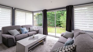 Ein modernes Wohnzimmer mit grauem Sofa, einem Holztisch und großen Fenstern mit Jalousien.
