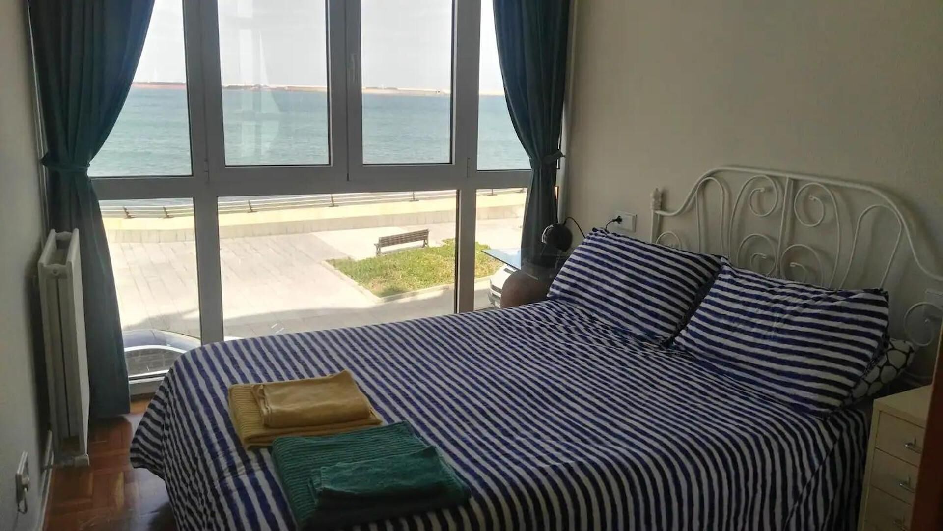 Una cama con ropa de cama a rayas, vista al mar a través de amplios ventanales y un cabecero de metal.
