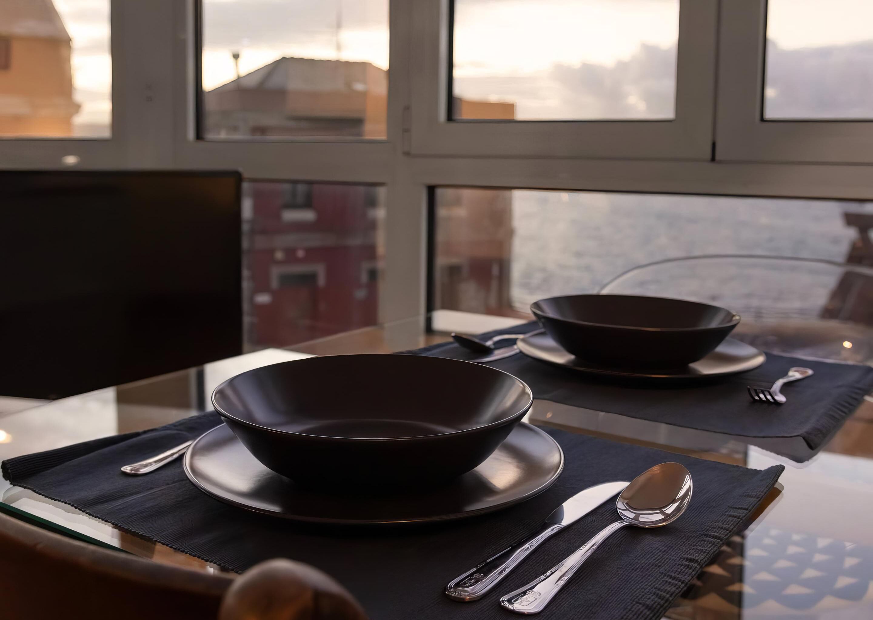 Una mesa de comedor con dos cuencos oscuros, un cuchillo y una cuchara sobre posavasos negros.