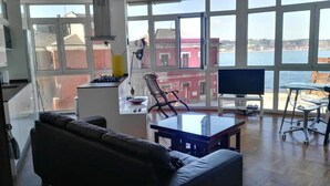 A spacious living area with a large window offering a view of the sea and buildings. There is a sofa, a coffee table, a TV, and a chair. - Seaviews-gijon-parking-2br-walk to Beach-wifi (Gijon)