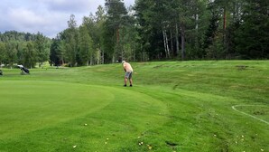 A golf course with a well-maintained green, a golfer in a white cap, and golf bags in the background. - Rosa's Stuga, a lovely house for two people in the middle of beautiful nature.   (Klässbol)