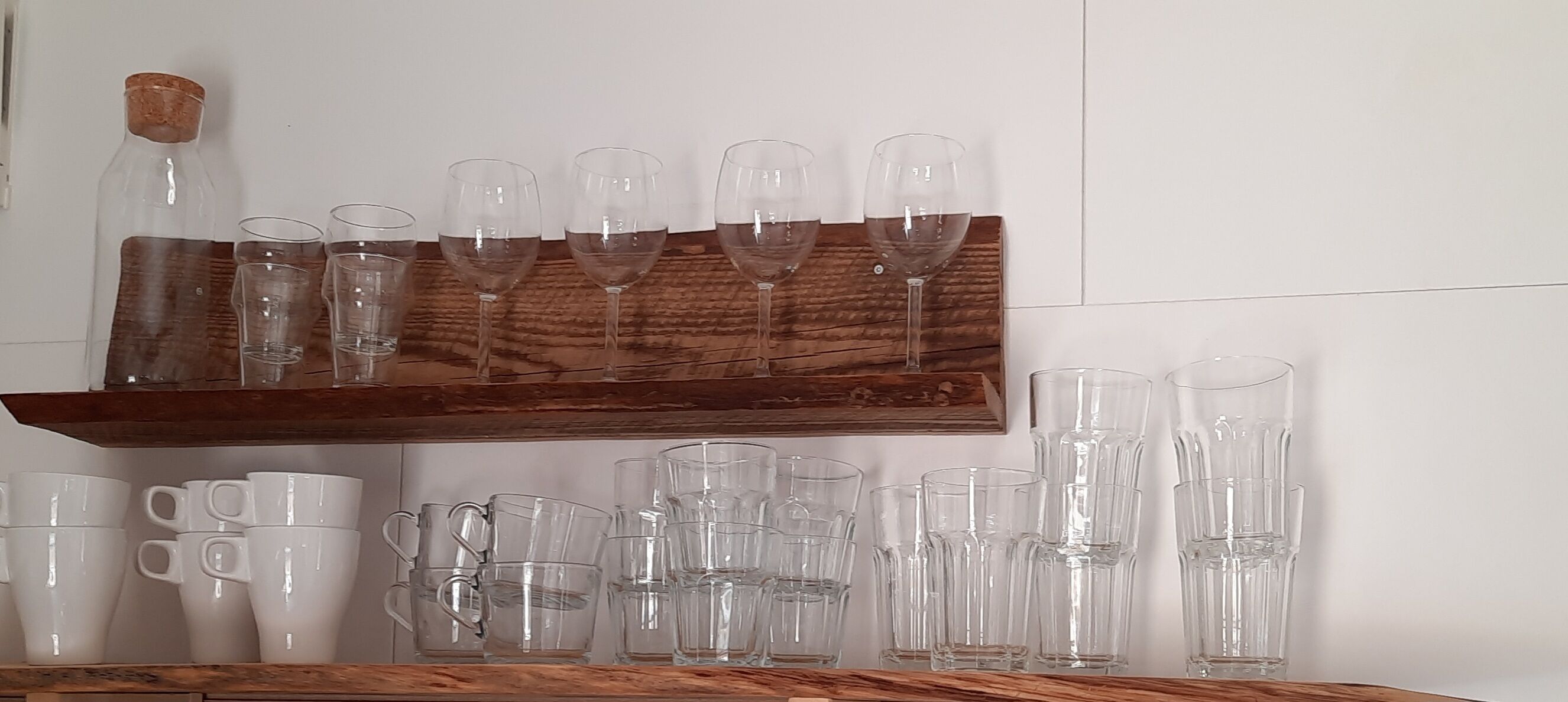 En hylla i trä med olika typer av glasvaror, inklusive vinglas och dricksglas, placerad mot en vit vägg.