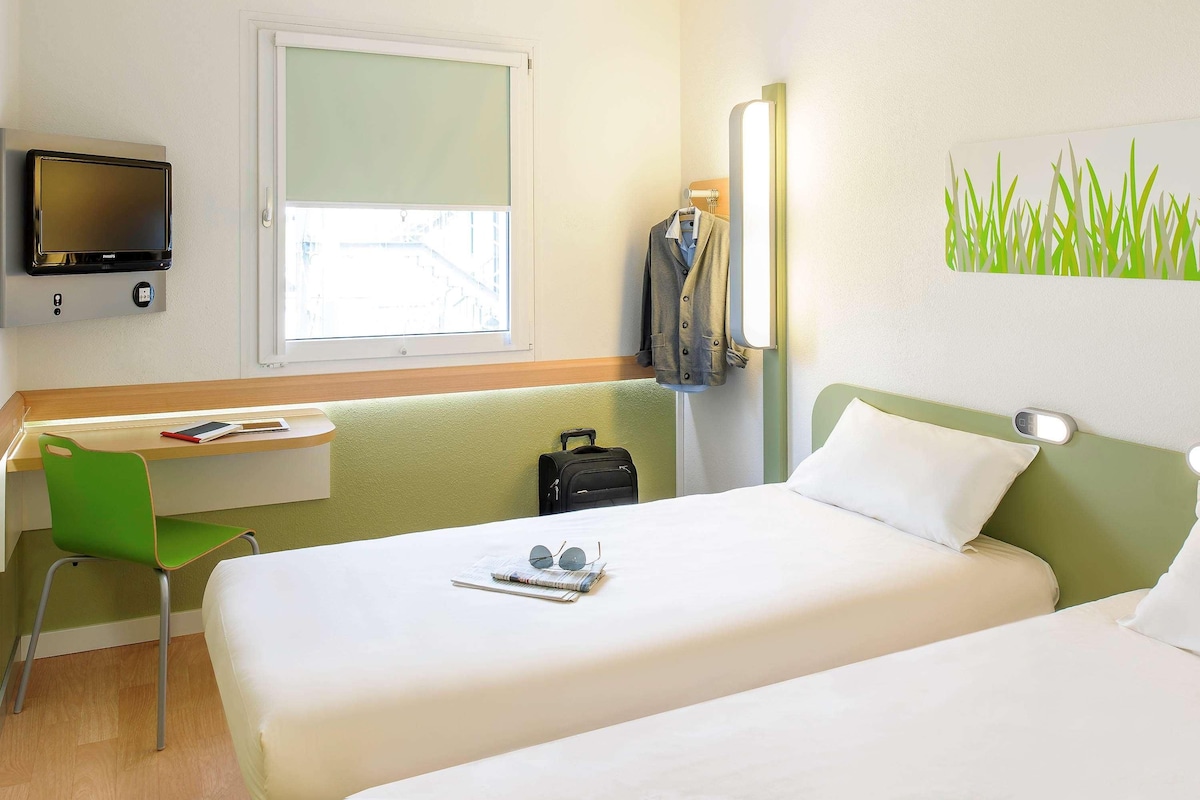 A hotel room with two beds, a desk, a chair, a TV, and a window.