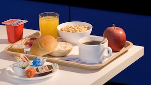 Bandeja de desayuno con un croissant, un muffin, un tazón de cereal, un vaso de jugo de naranja, una taza de café, una manzana roja, un paquete de Nutella, un envase de yogur y una taza roja.