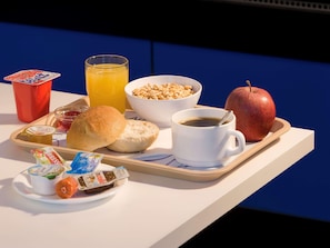 A breakfast tray with a croissant, a muffin, a bowl of cereal, a glass of orange juice, a cup of coffee, a red apple, a packet of Nutella, a packet of yogurt, and a red cup. - B&B Hotel Berlin Genshagen-nord (Ludwigsfelde)