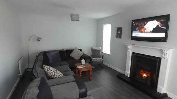 A living room with a grey sofa, a small table, a chair, a fireplace, and a TV mounted on the wall. - Tranquil Retreat Close to the Coast (Coleraine)