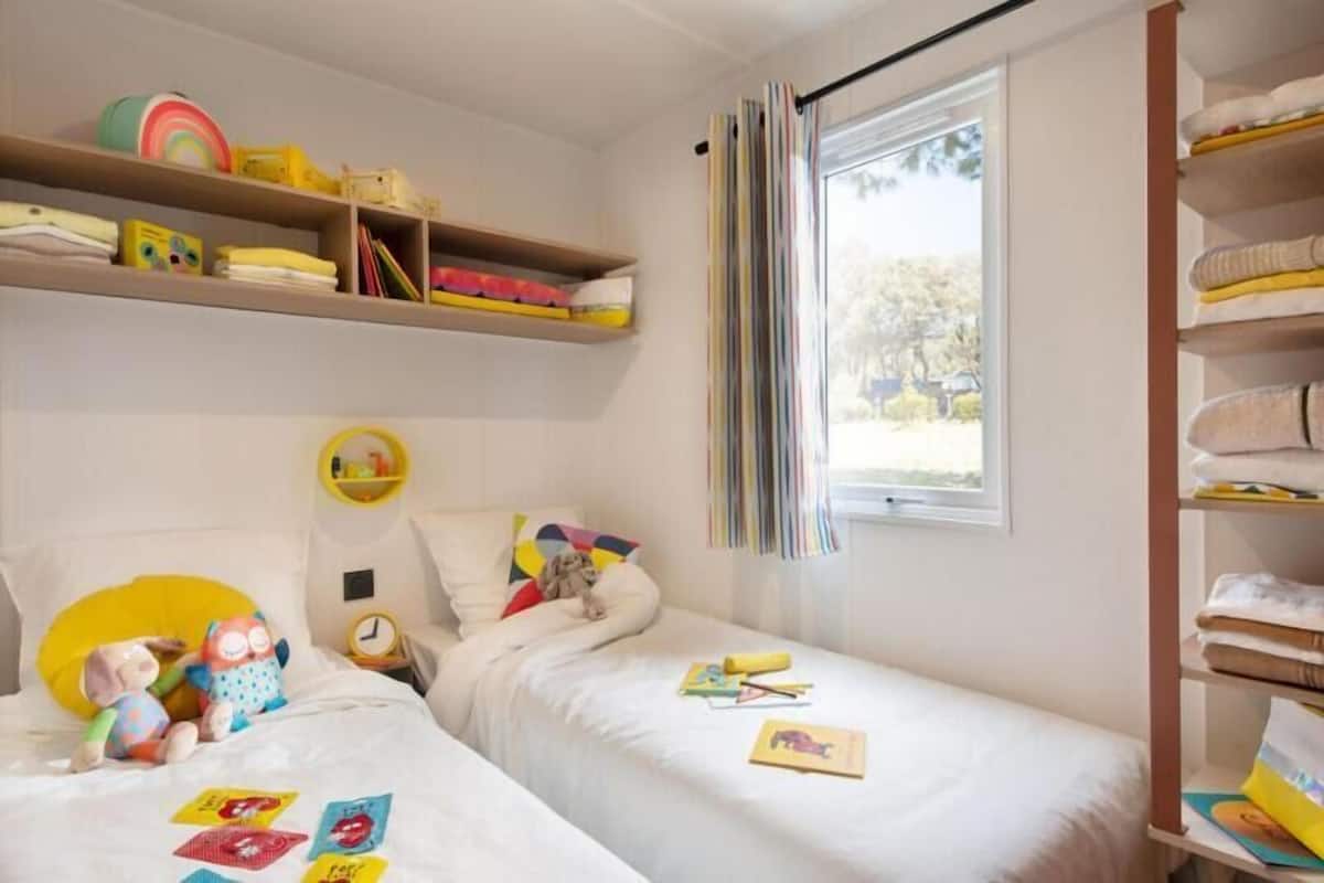 A child's bedroom with two beds, a shelf with toys, and a window with curtains.