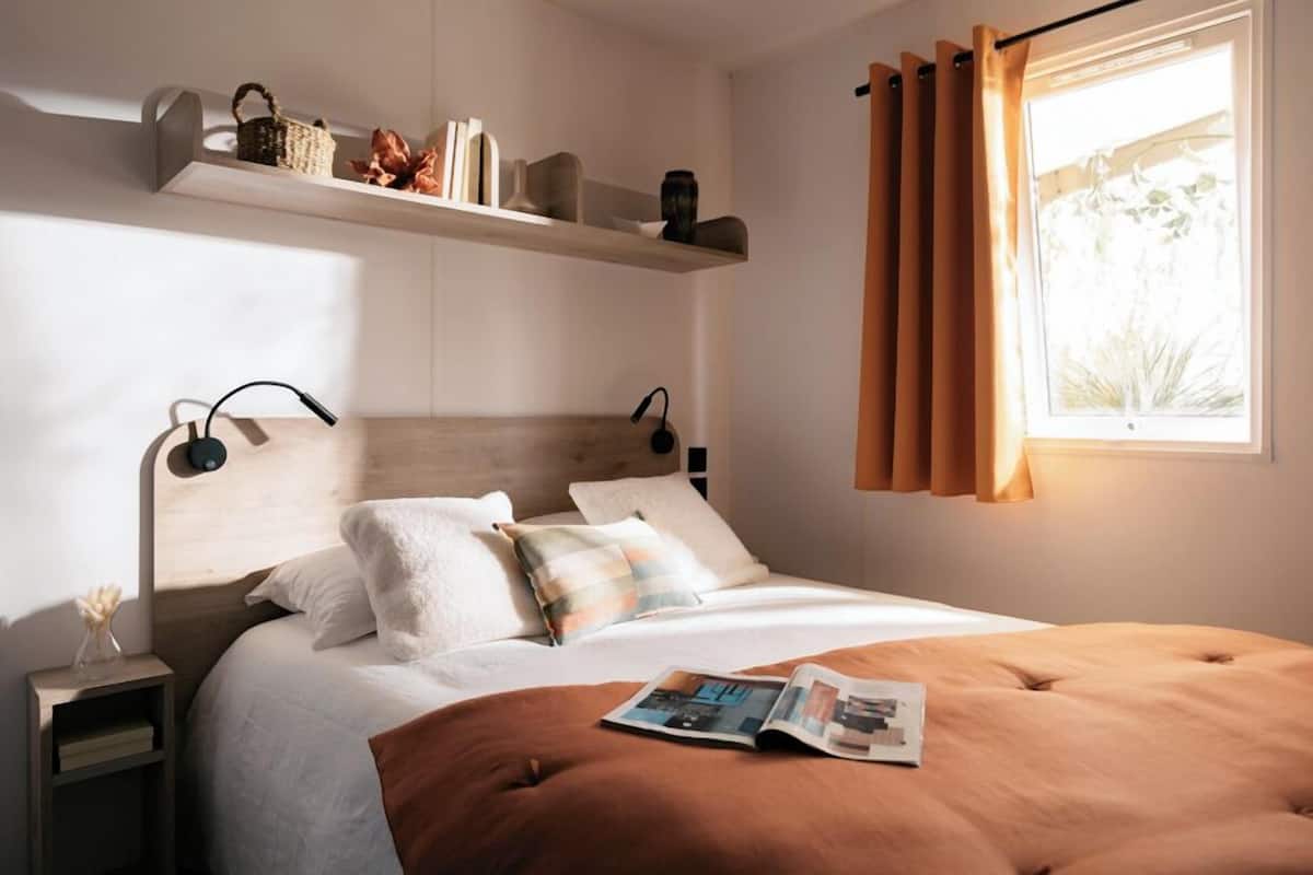 A neatly arranged bedroom with a wooden headboard, a bedside table with a lamp, a shelf with decorative items, and a window with curtains.