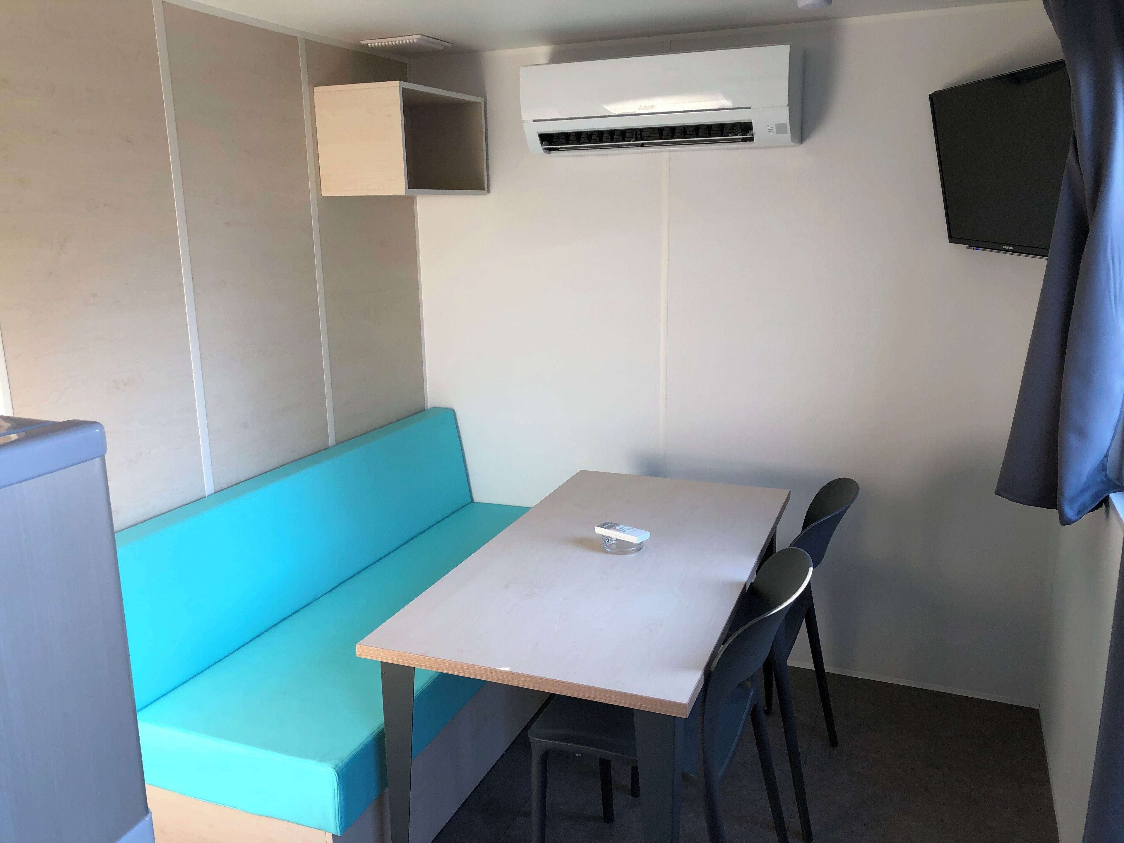 A small, modern room with a teal bench, a wooden table, and two chairs. An air conditioning unit is mounted on the wall.
