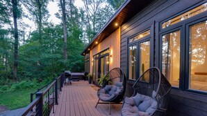 A wooden deck with two wicker chairs, a barbecue grill, and a large window overlooking a forested area. - Woodland Chalet  Hot Tub  Fire Pit  Grill (Quarryville)