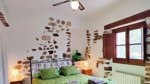 A bedroom with a stone wall, a bed with green bedding, a nightstand, a lamp, and a window with wooden shutters. - Beautiful views of the Sierra de Grazalema, ideal to enjoy and disconnect. (Montecorto)