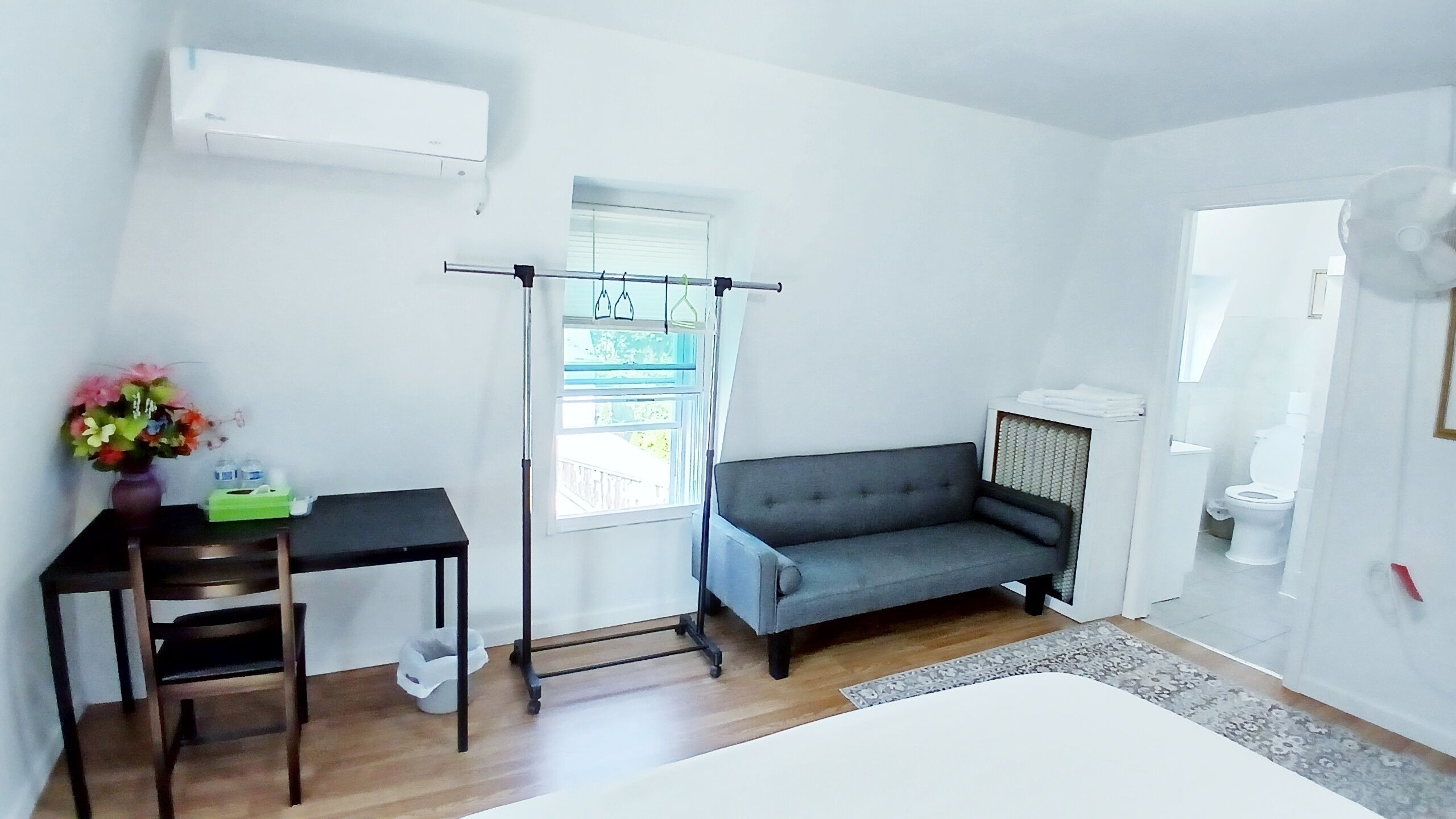 Een compacte kamer met een grijze bank, een klein tafeltje met een stoel, een raam met uitzicht en een witte airconditioning op de muur.