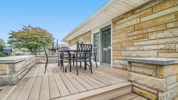 Une terrasse en bois avec un muret en pierre, une table avec des chaises et une porte-fenêtre coulissante.