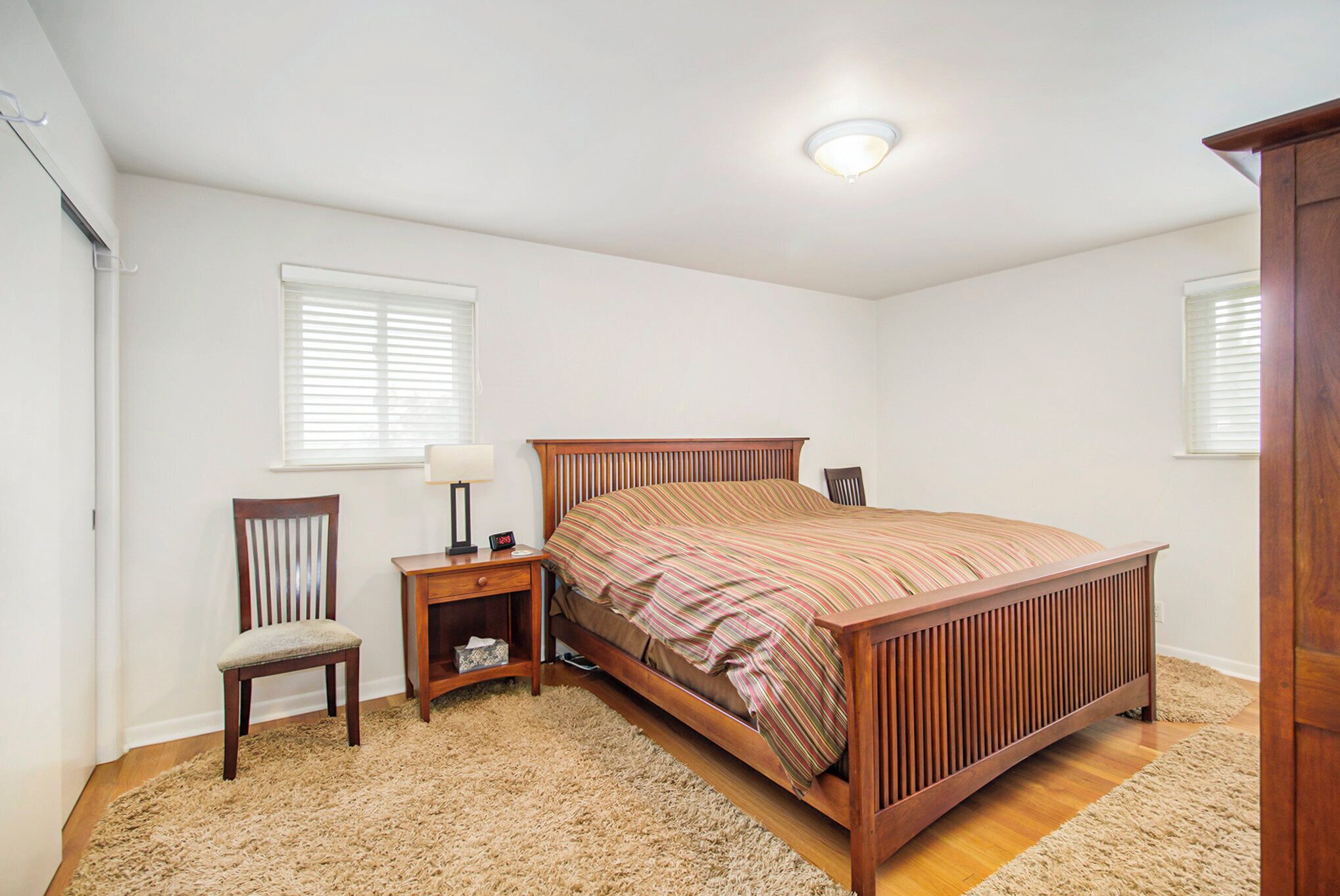 Een slaapkamer met een houten bed, een nachtkastje, een stoel en een raam met jaloezieën.