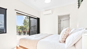 A bedroom with a large bed, a sliding glass door leading to a balcony with potted plants, and an air conditioning unit on the wall. - Your Ultimate Escape in Mooroobool- Luxury Tropical Retreat with Pool & Stunning Views – Perfect for Families & Groups! (Mooroobool)
