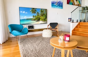 Una sala de estar con una televisión de pantalla plana, una silla azul, un taburete blanco, una mesa de centro de madera y una escalera.