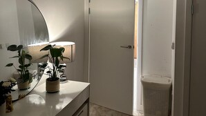 A bathroom with a white vanity, a large mirror, a potted plant, and a trash bin. - Cosy Thameside Retreat in North Greenwich (London)