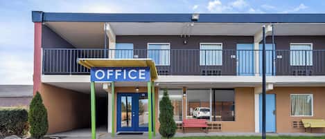 Un edificio de dos pisos con un letrero que dice "OFFICE" sobre la entrada. Tiene dos ventanas en la planta baja y un balcón con barandillas.