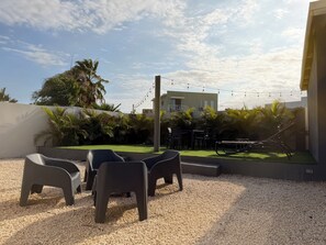 Een buitenruimte met stoelen en tafels, omgeven door begroeiing en een gebouw op de achtergrond.