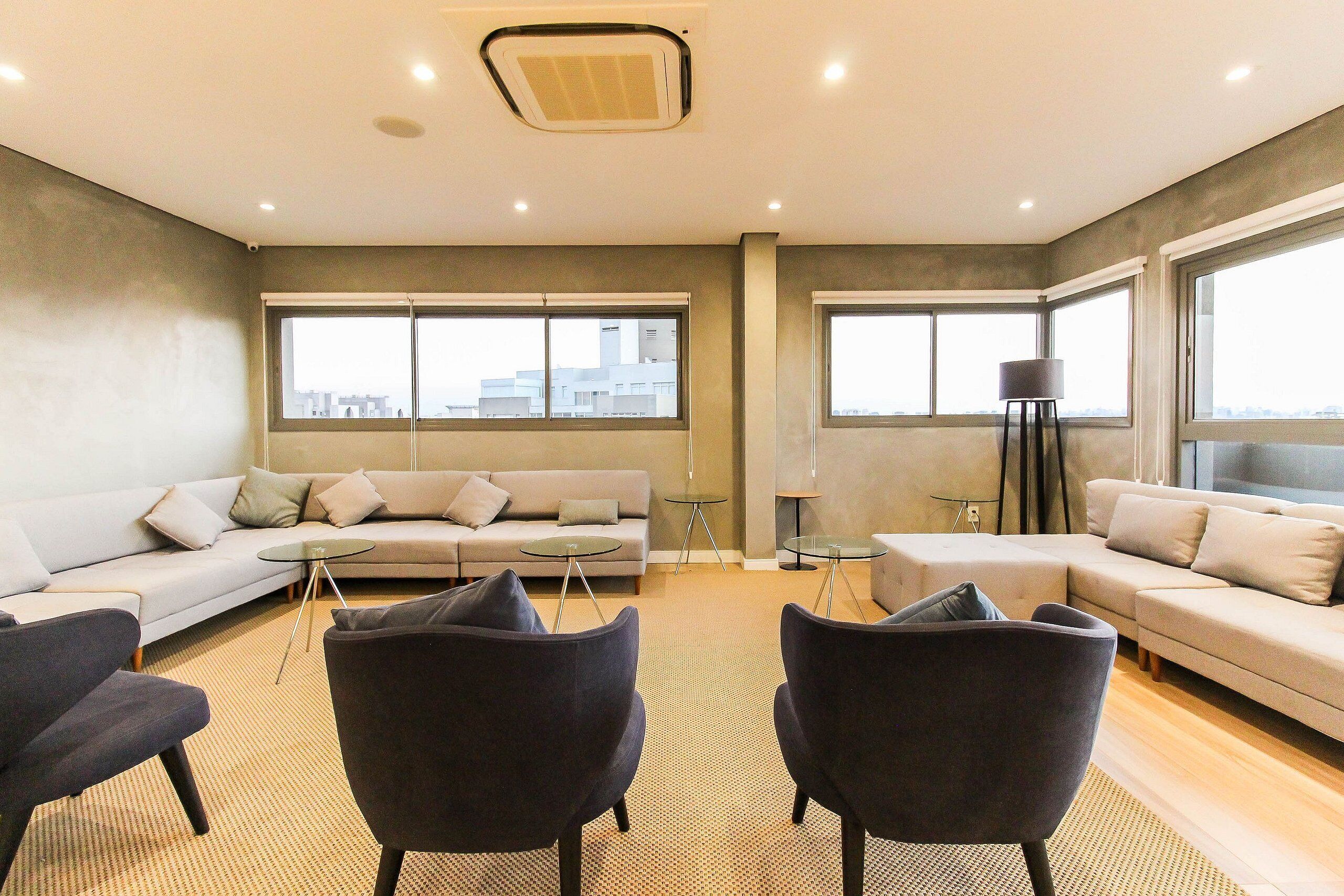 Una sala de estar moderna con un sofá, dos sillones, una mesa de centro y una lámpara de pie. Amplias ventanas ofrecen vistas del paisaje urbano.