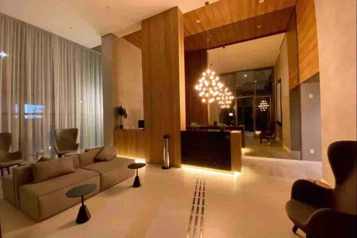 A modern living room with a sofa, coffee table, and chairs. There is a reception desk and a chandelier in the background.