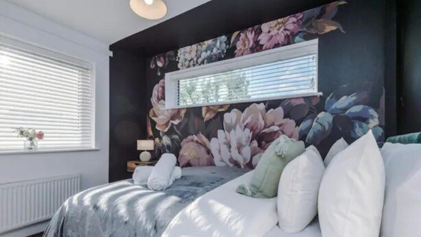 A bedroom with a large bed, a window with blinds, a floral wall mural, and a nightstand with a lamp. - Manchester Oasis Retreat - 4 Bedroom House (Manchester)