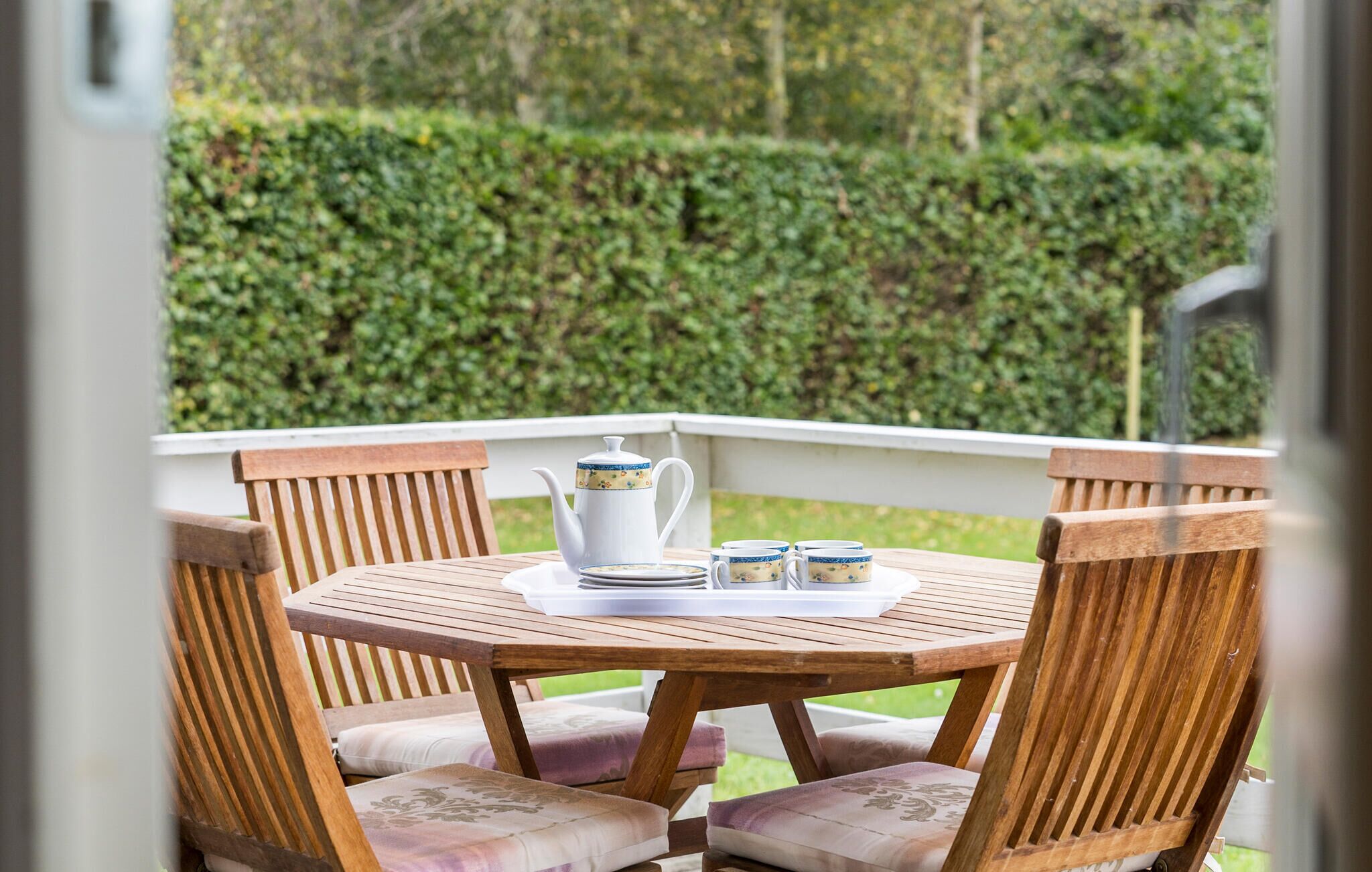 Een houten tafel met een witte theepot, kopjes en schotels op een balkon met houten stoelen.