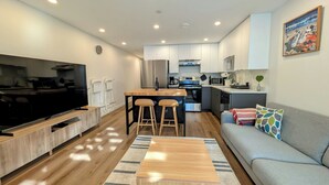 A modern kitchen with a central island, a dining area with a wooden table and chairs, and a grey sofa with colorful cushions. - Bright 2-Bed Ground Suite | Steps to PNE & Transit (Vancouver)