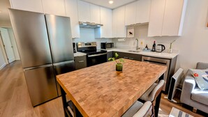 A modern kitchen with stainless steel appliances, a wooden dining table, and a grey sofa. - Bright 2-Bed Ground Suite | Steps to PNE & Transit (Vancouver)