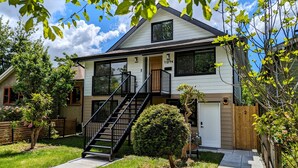 A two-story house with a front entrance, a visible number '1-2794', a black staircase leading to the entrance, and a small front yard with a bush and a wooden fence. - Bright 2-Bed Ground Suite | Steps to PNE & Transit (Vancouver)